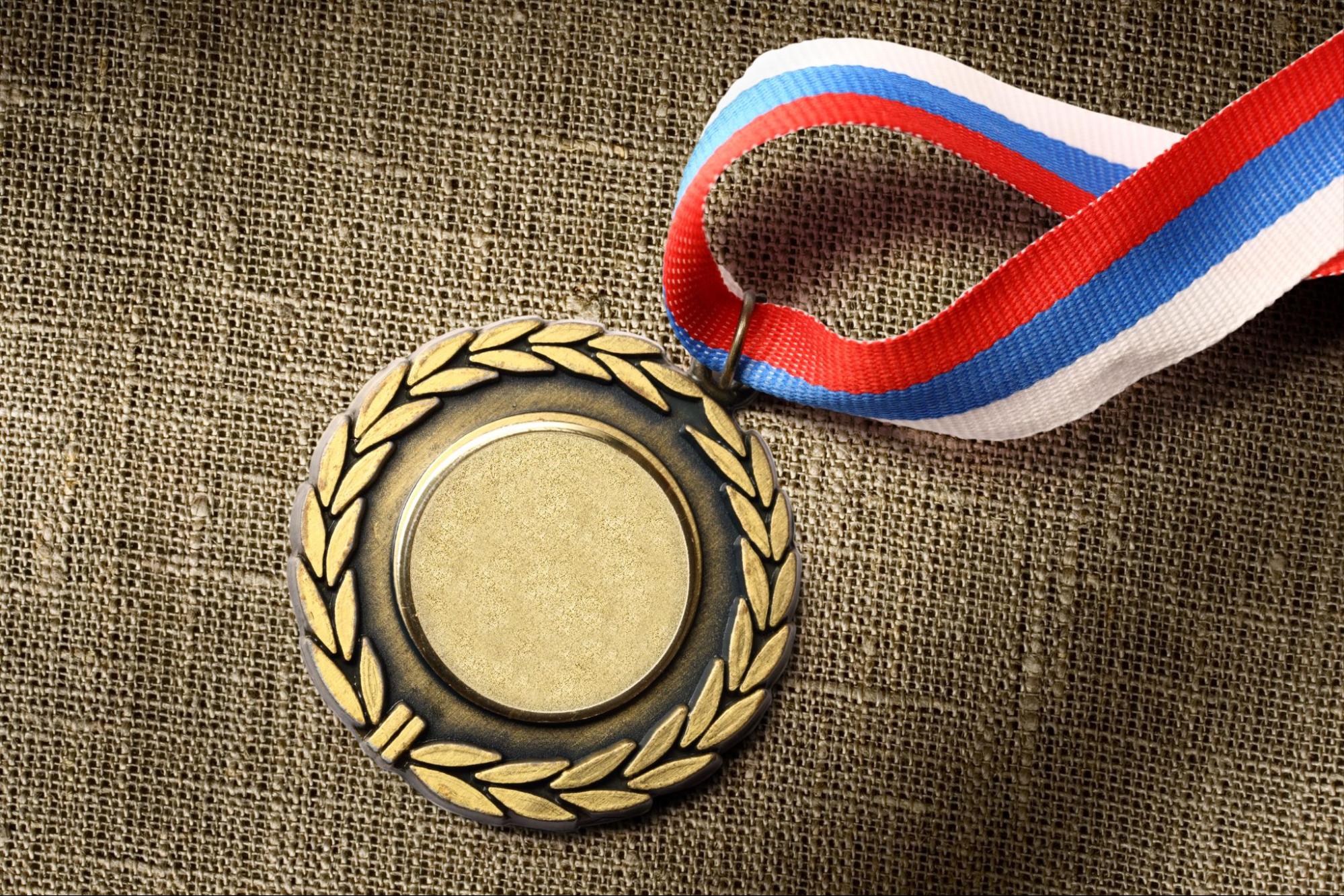 A gold medal with a laurel wreath design, placed on a textured fabric background. The medal is attached to a ribbon with red, white, and blue stripes. The center of the medal is blank, allowing for customization or engraving.